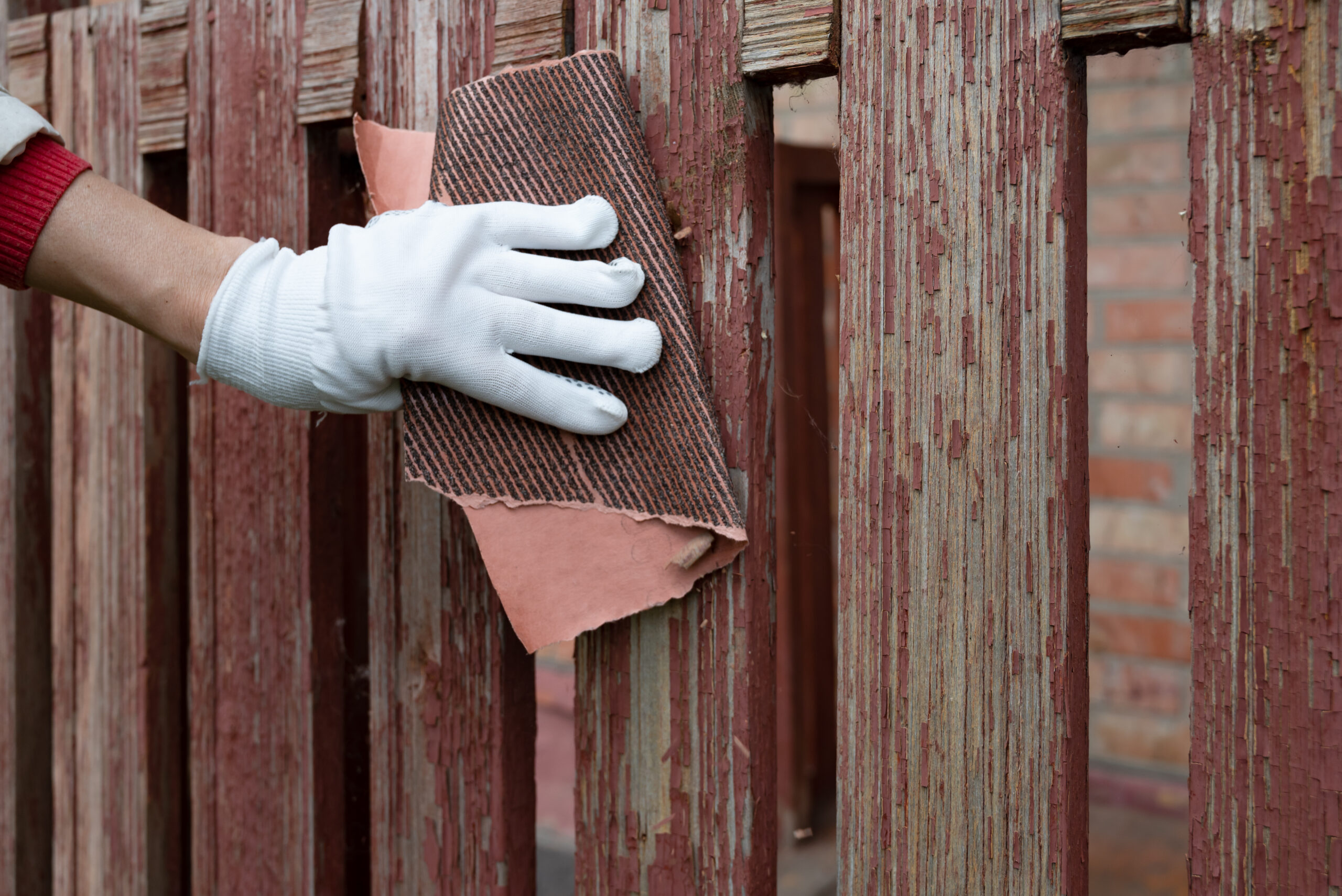 A person wearing a white glove is sanding a weathered wooden fence in the late afternoon. The fence shows signs of paint peeling and wear, indicating a restoration project.