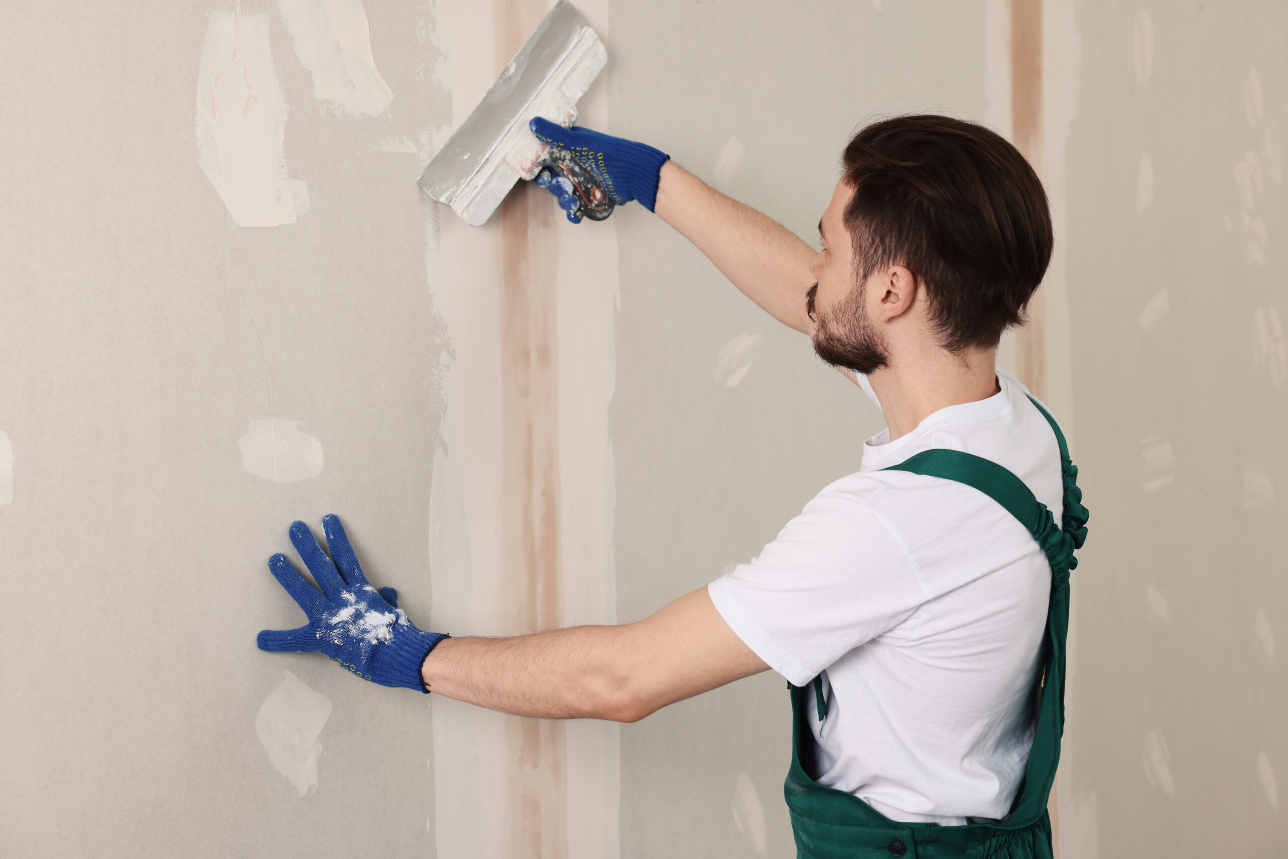Worker in uniform plastering wall with putty knife indoors